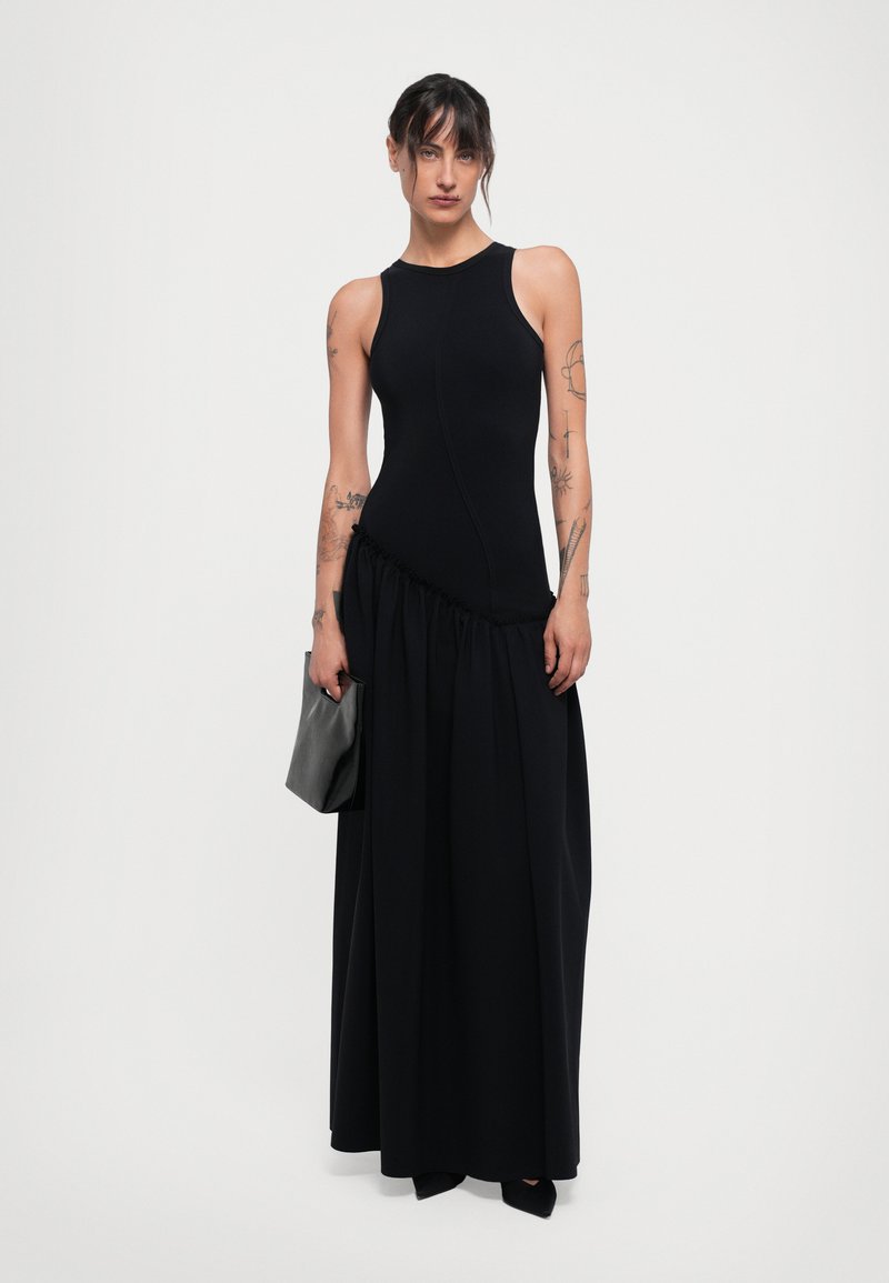 Woman with tattoos wearing a sleeveless black dress with asymmetrical gathered waist, holding a small black leather clutch and wearing black shoes.