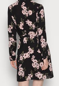 Abito nero a fiori con maniche lunghe, vita cintata e orlo a lunghezza ginocchio. Il tessuto ha una consistenza morbida con grandi fiori rosa e bianchi.