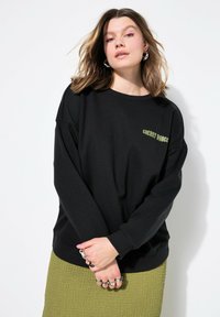 Felpa oversize nera con testo 'CHERRY DANCE' in verde. Presenta un collo rotondo e una texture morbida. Abbinata a una gonna verde con texture.