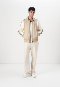 Beige Bomberjacke mit Reißverschluss, grünen und weißen Ärmeln, elastischen Bündchen und einem Logo-Patch auf der Brust. Kombiniert mit cremefarbenen Hosen und weißen Sneakers.