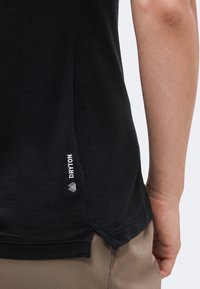 Κοντινό πλάνο στο πλάι του σώματος ενός ατόμου που φοράει μαύρο υφασμάτινο πουκάμισο με την ετικέτα «DRYTON» και μπεζ παντελόνια, με ορατό το δεξί του χέρι.