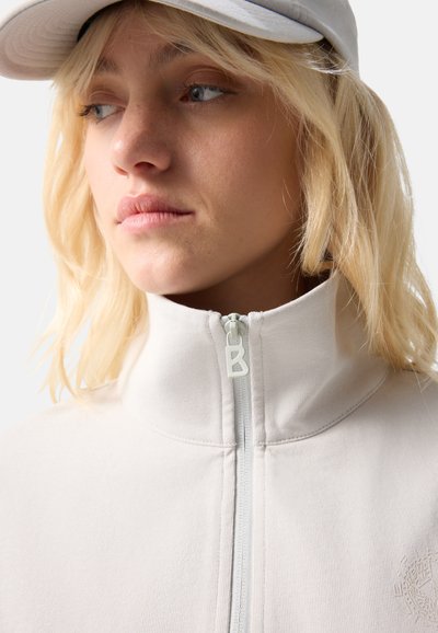 Young woman with blonde hair wears light gray cap and zipped high-collar cream jacket, looking to the side on a plain background.