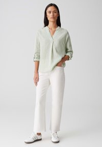 Camicia verde chiaro a maniche lunghe, con colletto e vestibilità rilassata, abbinata a pantaloni bianchi a gamba dritta e sneakers bianche.