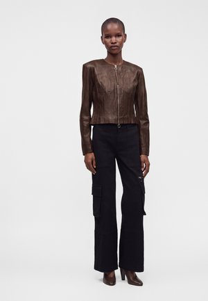 Personne portant une veste en cuir marron ajustée, un pantalon cargo noir à jambes larges et des chaussures marron à talons, debout devant un fond clair uni.