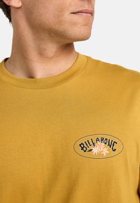 Gul bomulds-t-shirt med et cirkulært sort og lyserødt logo med blomster og teksten "BILLABONG". Klassisk rund hals, korte ærmer.