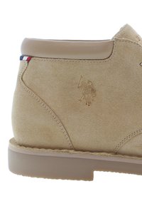Botín de ante beige con un suave cuello en tono miel, detalles de costura y un logotipo grabado. Presenta una suela de goma y un tacón ligero.