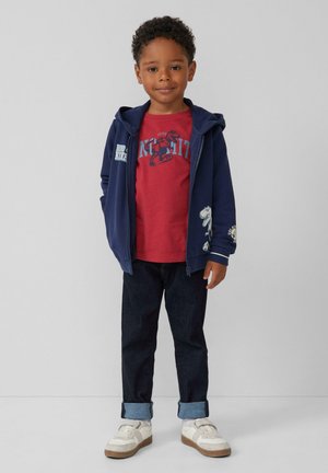 Marineblauwe rits hoodie met witte accenten, rode grafische T-shirt, donkere denim jeans en witte sneakers met bruine zolen.