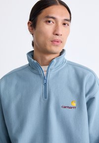 Sudadera de forro polar de cremallera azul claro con cuello alto, que presenta un logo de Carhartt bordado en amarillo y rojo en el pecho. Textura suave.