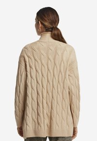 Beige kabelstickad cardigan med hög krage, som har texturerade vågmönster och ribbat avslut. Långa ärmar och avslappnad passform.