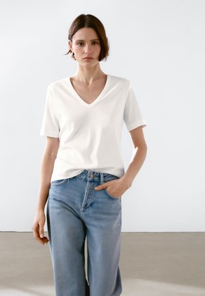 Donna con i capelli corti castani che indossa una maglietta bianca a V e maniche corte e jeans azzurri chiari, in piedi con una mano in tasca.