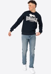 Navy crew neck sweatshirt with white lion graphic and "LONSDALE LONDON" text. Paired with light blue jeans and white sneakers.