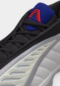 Κοντινό πλάνο μαύρου, λευκού και μπλε αθλητικού παπουτσιού με υφή διχτυού, μαύρα κορδόνια και κόκκινο τριγωνικό σχέδιο στη μπλε γλώσσα.