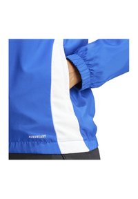 Blauwe windjack met witte accenten, elastische manchetten en AeroReady-branding, met een zijzak met een gladde, gestructureerde afwerking.