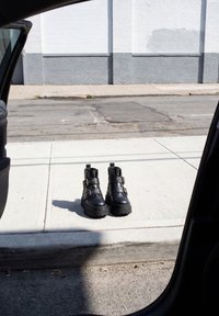 Paire de bottes de combat en cuir noir avec boucles, posées sur un trottoir ensoleillé, vues de l’intérieur d’une voiture avec la porte ouverte.