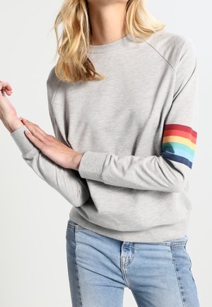 Osoba ubrana w jasnoszary sweter z tęczowym paskiem na lewym rękawie oraz wyblakłe niebieskie dżinsy, pozująca z założonymi skrzyżowanymi ramionami.
