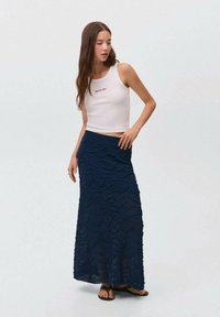 Top court blanc avec texte rouge, associé à une longue jupe tricot bleu marine avec des motifs en vagues. Des sandales complètent la tenue.