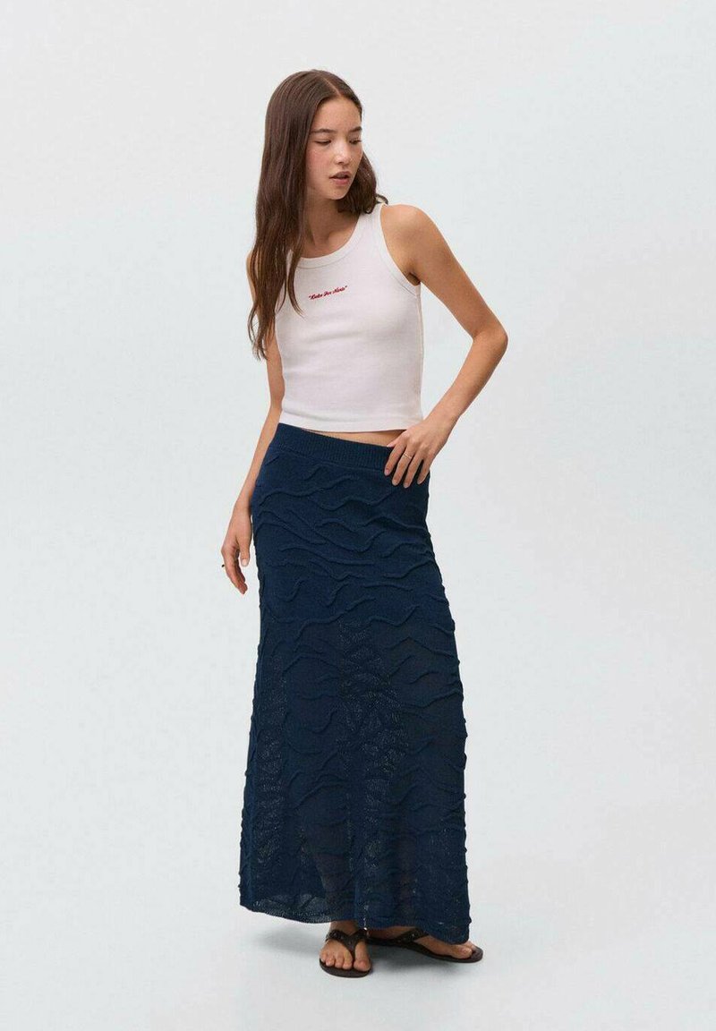 Top court blanc avec texte rouge, associé à une longue jupe tricot bleu marine avec des motifs en vagues. Des sandales complètent la tenue.