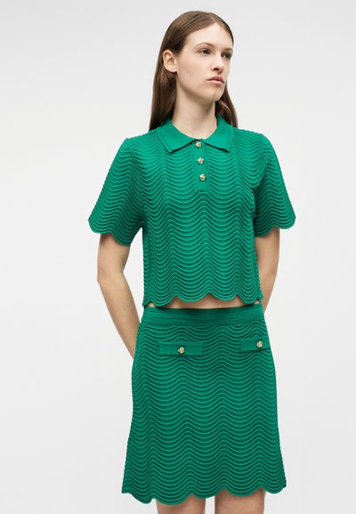 Jeune femme portant un polo en tricot vert texturé et un ensemble jupe assorti avec des bords festonnés et des détails de boutons dorés.