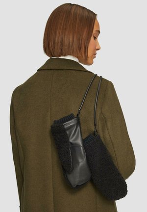 Femme aux cheveux bruns courts portant un manteau vert olive, portant des moufles en polaire et cuir noires reliées par une sangle sur son épaule.