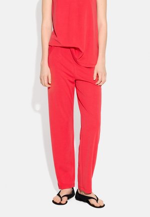 Osoba ubrana w czerwoną bluzkę bez rękawów i dopasowane spodnie z czarnymi sandałami, stojąca przed jednolitym jasnym tłem.