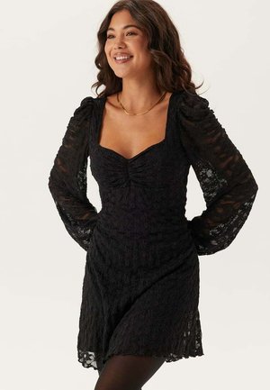 Svart spetsklänning med fyrkantig halsringning, rynkad front och långa puffärmar. Har strukturerad tyg och en utsvängd kjoldesign.