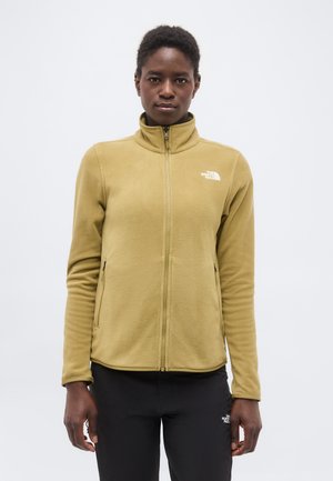 Persona in piedi, rivolta in avanti, che indossa una giacca in pile beige con cerniera e pantaloni neri, entrambi con il logo The North Face.