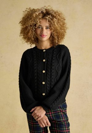 Schwarze Strickjacke mit goldenen Knöpfen, die ein Zopfmuster hat und weite Ärmel bietet. Das Modell trägt sie über einer Hose mit Karomuster.