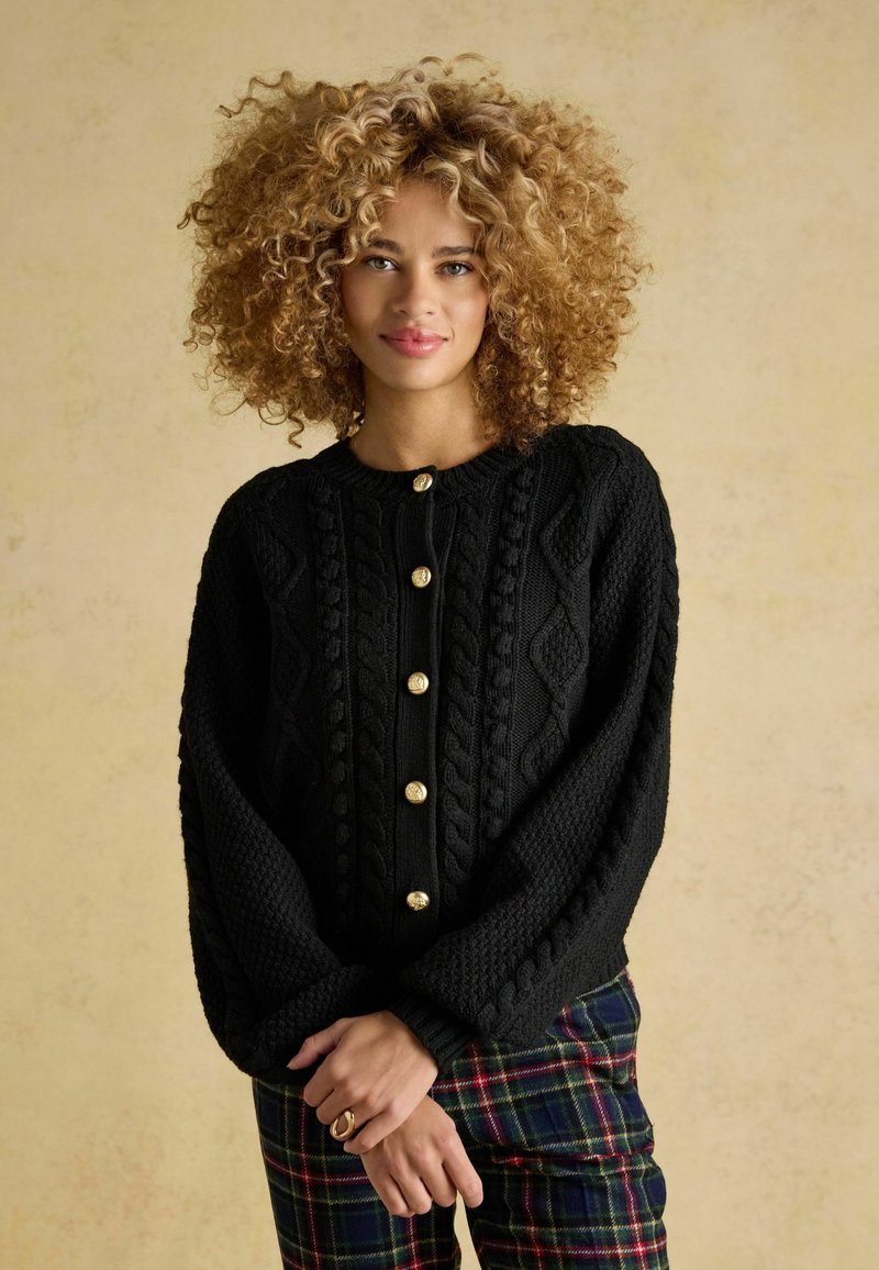 Czarny sweter robiony na drutach z złotymi guzikami, z splotem w warkocze i szerokimi rękawami. Model nosi go na spodniach w kratę.