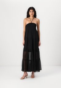 Fekete, pánt nélküli maxi ruha smocked bodiccel, réteges, áttetsző anyaggal és lebegő fazonnal. A modellen meztelen, pántos magassarkú cipők vannak.
