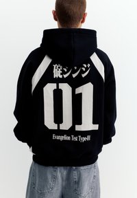 Person iført sort hættetrøje med hvidt nummer "01", japanske tegn og teksten "Evangelion Test Type-01" på ryggen, kombineret med grå jeans.