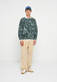 Adīta zaļi-zila džemperis ar teksturētu rakstu, apvienots ar bēšiem platām biksēm un baltām sporta kedām. Zils cepure noslēdz apģērbu.