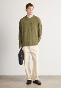 Maglione lavorato a maglia verde oliva con una finitura testurizzata, abbinato a pantaloni beige chiari a gamba larga e scarpe in pelle nere, mentre tiene una borsa nera.