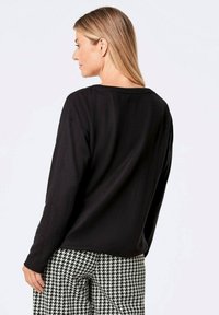 Schwarzes langärmliges Oberteil mit einem leicht bootförmigen Ausschnitt, aus einem glatten, strukturierten Stoff; kombiniert mit hochkontrastierenden Hahnentritt-Musterhosen.