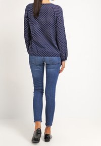 Marinblus med cirkulära mönster, kombinerad med åtsittande blå jeans och svarta lackskor. Har långa ärmar och rund halsringning.