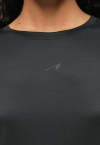 Κοντινή λήψη ενός ατόμου που φοράει ένα σκούρο γκρι μπλουζάκι με στρογγυλή λαιμόκοψη, με ένα μικρό, διακριτικό λογότυπο κοντά στο κέντρο του λαιμού.