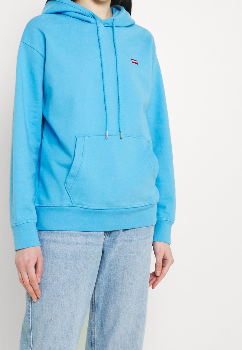 Personne portant un sweat à capuche bleu clair avec poche avant et un jean bleu clair, debout devant un fond blanc uni.