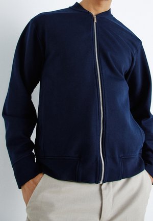Homme portant une veste zippée bleu marine avec des poignets côtelés et un pantalon beige, mains dans les poches, sur un fond uni.