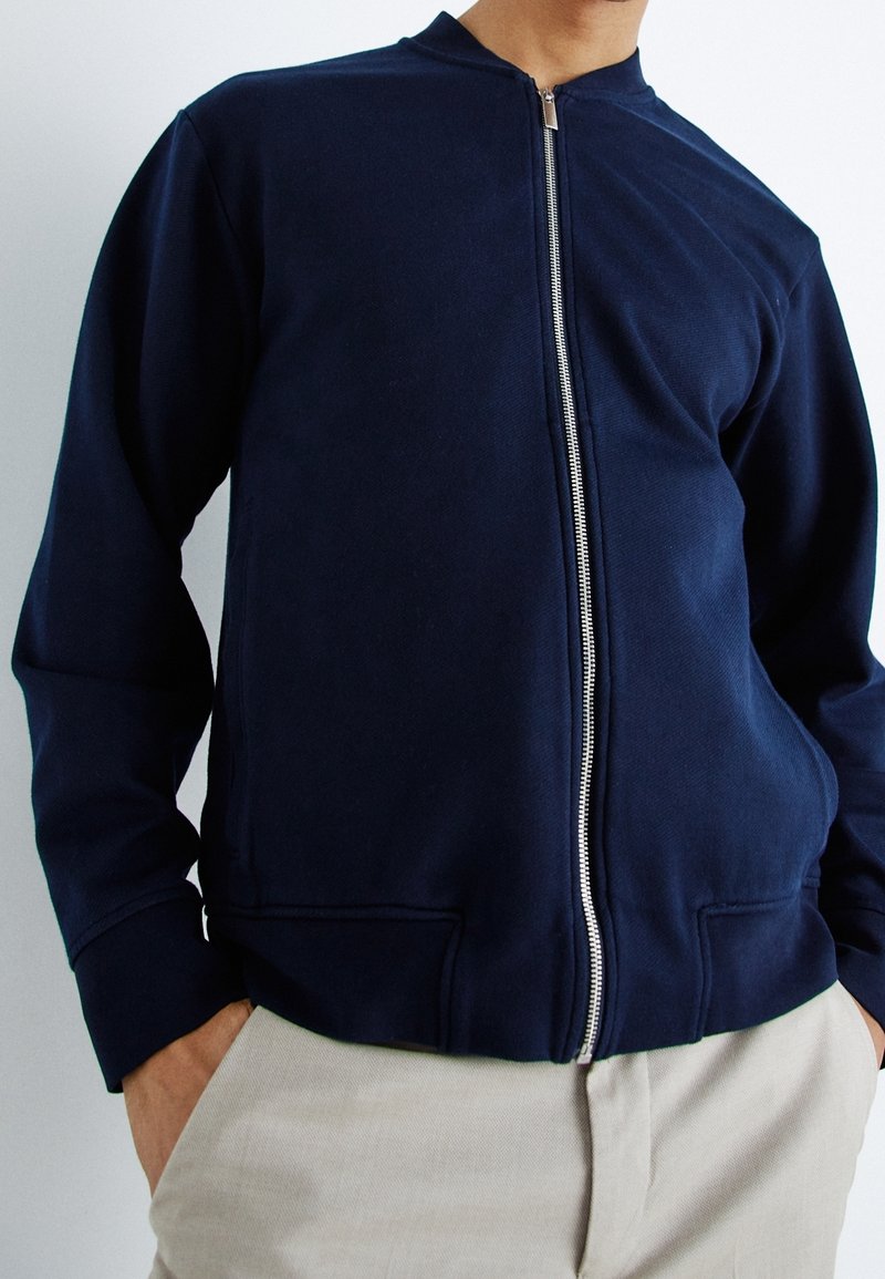 Homme portant une veste zippée bleu marine avec des poignets côtelés et un pantalon beige, mains dans les poches, sur un fond uni.