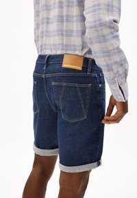 Donkerblauwe denim shorts met omgeslagen zoom, voorzien van een leren label met de tekst "ARMEDANGELS" en een vijf-pocketontwerp.