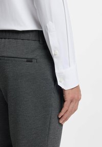 Man draagt een wit shirt met lange mouwen en donkergrijze, gestructureerde broek, met hand ontspannen naast de heup tegen een witte achtergrond.