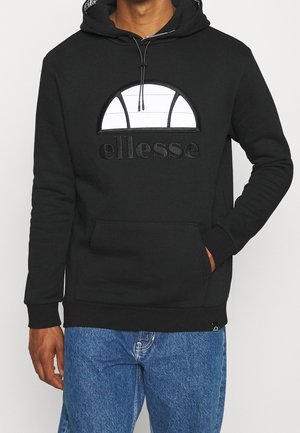 Czarna bluza z kapturem ellesse z białym logo w kształcie półkola na piersi, przednia kieszeń kangurka, noszona z niebieskimi jeansami, ręka w kieszeni, stojąca na białym tle.