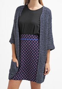 Lång, öppen cardigan i mörkgrått stickat med ett texturerat mönster. Tillsammans med en svart topp och en mönstrad lila kjol med diamanter.