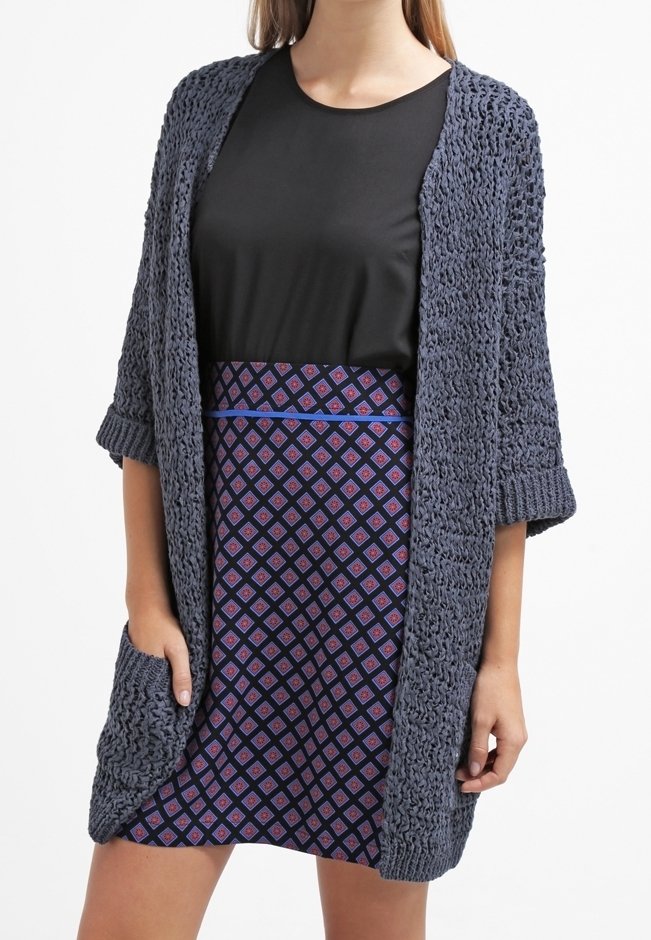 Lång, öppen cardigan i mörkgrått stickat med ett texturerat mönster. Tillsammans med en svart topp och en mönstrad lila kjol med diamanter.