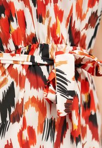 Kangas vestido con un patrón abstracto multicolor en rojo, naranja, negro y crema. Presenta una cintura ajustada y un detalle de cinturón atado.