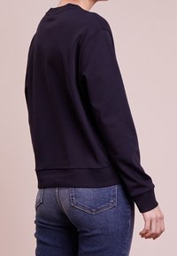 Granatowa bluza z okrągłym dekoltem, długimi rękawami oraz prążkowanymi mankietami i dołem, zestawiona z dopasowanymi niebieskimi jeansami, przedstawiona od tyłu.