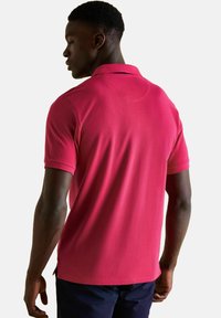 Muž v jasně růžové polokošili s krátkým rukávem, zobrazený zezadu na bílém pozadí.