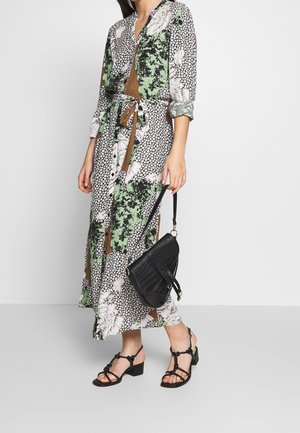 Kobieta ubrana w długą, kwiatową sukienkę z motywem patchworku i podwiniętymi rękawami, trzymająca czarną torebkę o fakturze oraz nosząca czarne sandały na słupku z paskami.