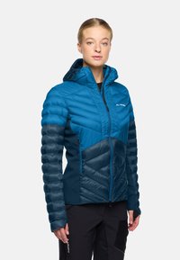 Donna che indossa una giacca a vento isolata blu e navy con cappuccio e cintura frontale, e pantaloni neri, in piedi davanti a uno sfondo bianco uniforme.
