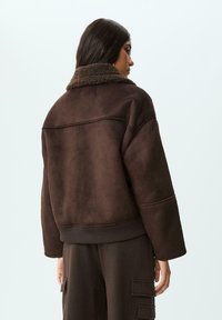 Giacca bomber in finta pelle scamosciata marrone con un morbido colletto in shearling, polsini a coste e vestibilità rilassata. Dettagli cucitura sulle spalle e sul retro.