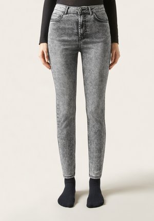 Mujer con vaqueros pitillo de talle alto en lavado ácido gris y calcetines negros, de pie frente a un fondo liso.